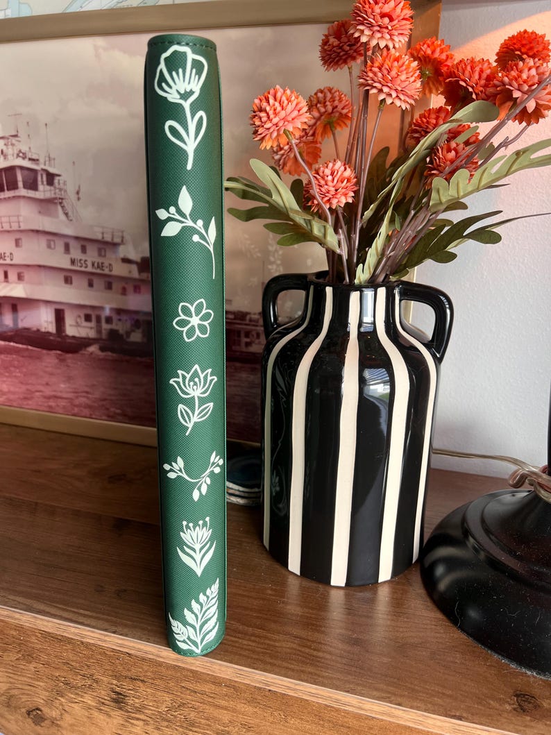 May include: A dark green vertical book spine featuring a series of white line-art botanical illustrations including various flowers, leaves, and sprigs arranged in a column along the center of the cover surface.