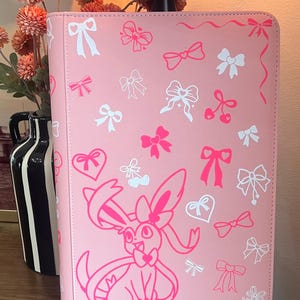 May include: A pink binder featuring a repeating pattern of white and hot pink bows, hearts, and cherry motifs. A large, stylized hot pink graphic of the character Sylveon is printed on the lower front, accented by additional bow and heart illustrations.