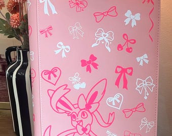 May include: A pink binder featuring a repeating pattern of white and hot pink bows, hearts, and cherry motifs. A large, stylized hot pink graphic of the character Sylveon is printed on the lower front, accented by additional bow and heart illustrations.