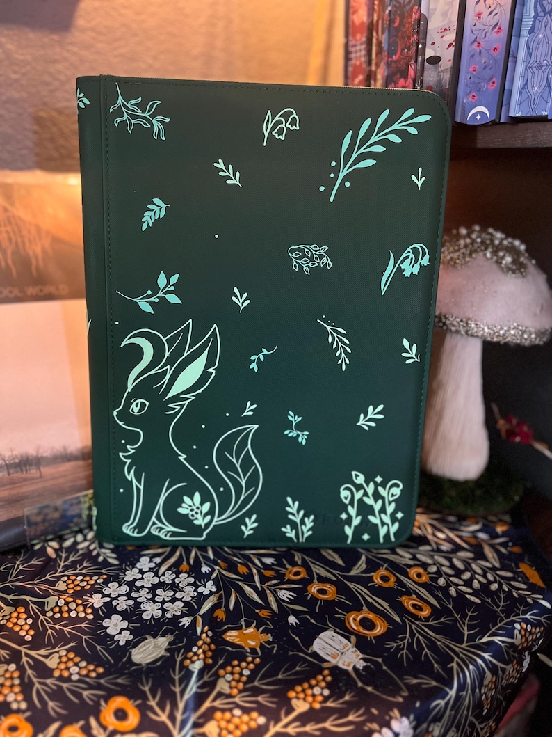 May include: A dark green rectangular notebook cover featuring a light mint green graphic of a stylized fox-like creature with large ears and a crescent moon shape on its head, surrounded by various small botanical leaf and flower illustrations.