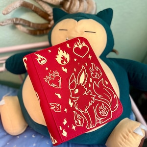 May include: A red book cover featuring a white illustration of a fox-like creature with flames, hearts, and fire symbols. The cover is closed and has a soft, textured appearance. The background includes a large plush toy.