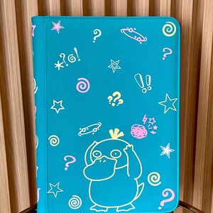 May include: A teal rectangular binder featuring a yellow outline of the character Psyduck at the bottom. The cover is decorated with various scattered graphic elements in pink, yellow, and white, including stars, question marks, spirals, and exclamation points.