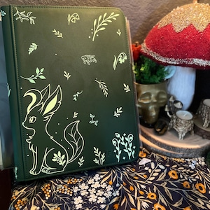 May include: A dark green binder featuring a light green graphic of a stylized fox-like creature surrounded by various botanical elements including leaves, sprigs, and small flowers printed across the front cover surface.