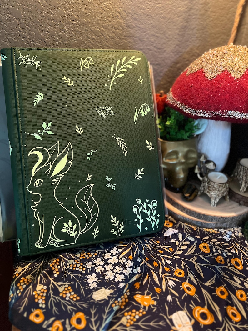 May include: A dark green binder featuring a light green graphic of a stylized fox-like creature surrounded by various botanical elements including leaves, sprigs, and small flowers printed across the front cover surface.