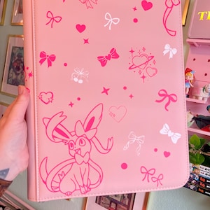 Puede incluir: Carpeta rosa claro con un patrón repetido de lazos, corazones y estrellas rosas y blancas. Una gran ilustración rosa de un personaje de Sylveon aparece en la parte delantera. La carpeta es sostenida por una persona.