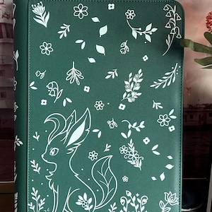 May include: A dark green rectangular binder featuring a white line art pattern of various small flowers, leaves, and a stylized fox-like creature with large ears and a bushy tail. The surface has a textured finish and a vertical spine on the left.