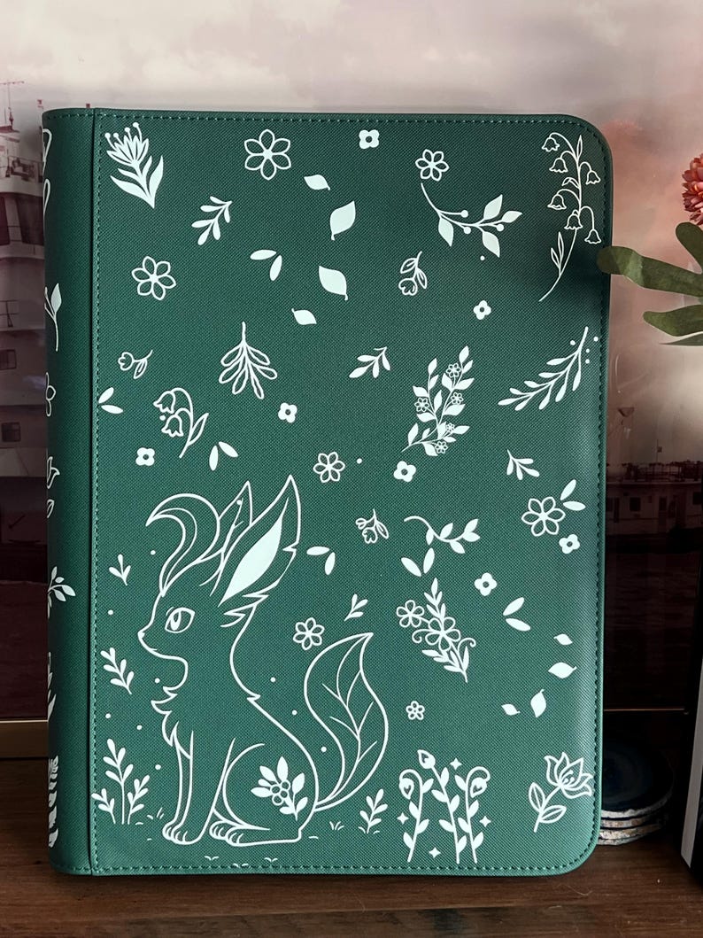 May include: A dark green rectangular binder featuring a white line art pattern of various small flowers, leaves, and a stylized fox-like creature with large ears and a bushy tail. The surface has a textured finish and a vertical spine on the left.