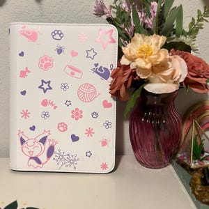May include: A white binder featuring a pink and purple cat-themed design. The pattern includes paw prints, stars, hearts, and cat-related icons. The binder is upright next to a vase of flowers.