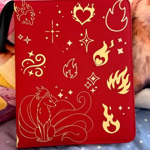May include: Red binder with a black handle featuring gold illustrations of flames, stars, and a nine-tailed fox. The binder has a smooth texture and a black handle for carrying.