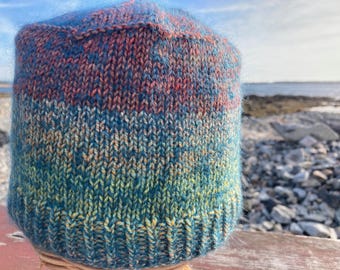 Op de afbeelding: Een handgebreide beanie-muts met een kleurverloop. De muts loopt van dieprood naar blauw- en groentinten. Gemaakt van zacht, getextureerd garen, ideaal voor koud weer. De muts ligt op een houten oppervlak.