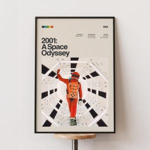 Puede incluir: Póster enmarcado de la película "2001: Una odisea del espacio". El póster tiene un fondo beige con el título de la película en letras negras grandes. Un astronauta con un traje espacial naranja camina por un pasillo blanco. El póster está en un marco negro.
