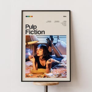 以下が含まれることがあります： 黒いフレームに入った映画「パルプ・フィクション」のポスター。ポスターには映画のタイトルと映画のシーンが描かれています。画像には、ベッドに横たわり、タバコと銃を持っている女性が写っています。ポスターは1994年製です。
