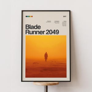 以下が含まれることがあります： 黒いフレームに入った映画「ブレードランナー2049」のポスター。ポスターには、太字のタイトルが、シルエットのあるオレンジと黄色の風景の上に表示されています。2017年が上部に表示されています。