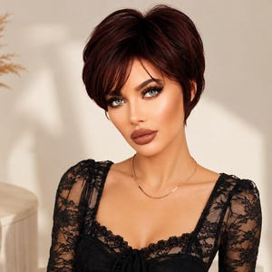 May include: A short, layered dark burgundy wig with side-swept bangs and a textured, voluminous crown. The hair is styled in a cropped cut that frames the face, featuring deep red and brown tones throughout the synthetic fibers of the hairpiece.