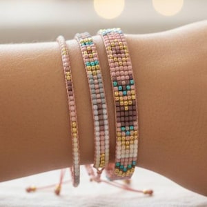 May include: Four beaded bracelets in shades of pink, gold, and teal are displayed on a person's arm. The bracelets have intricate patterns and textures, with a delicate, handcrafted appearance. The background is softly blurred with bokeh lights.