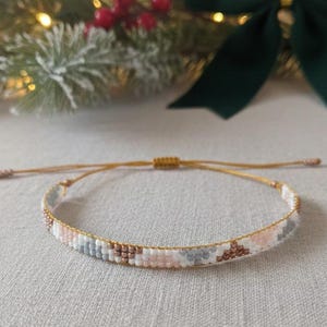 Puede incluir: Una pulsera tejida con un patrón de cuentas blancas, rosas, grises y bronce. La pulsera está en un cordón dorado. El fondo incluye un lazo verde, ramas de pino y luces de cadena.