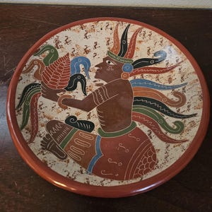 May include: A round ceramic plate with a brown rim and a cream-colored center. The plate features a detailed illustration of a figure holding a bird, with colorful accents in red, blue, green, and black. The design has an ancient, cultural theme.