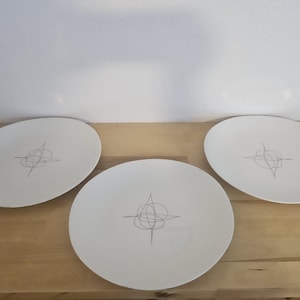 May include: Three round white ceramic plates, each featuring a centered, thin, dark gray line drawing of an abstract, geometric, star-like pattern composed of intersecting loops and straight lines, arranged on a light-colored wooden surface.