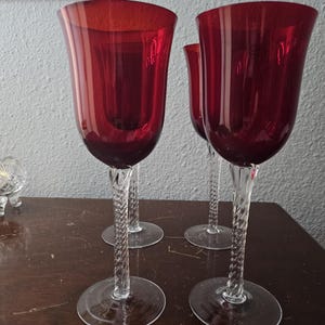 May include: A set of four wine glasses featuring deep red bowls and clear, twisted glass stems. Each glass has a wide, circular base and a smooth, flared rim. The stems have a distinct spiral pattern that connects the base to the colored bowl.