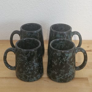 May include: A set of four ceramic mugs featuring a dark black base color with a mottled, speckled pattern in shades of teal and grey. Each mug has a cylindrical body, a rounded handle on the side, and a smooth, glossy finish throughout the exterior.