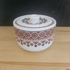 May include: A white ceramic sugar bowl with a lid, featuring a repeating geometric pattern in shades of brown, black, and tan. The bowl has a cylindrical shape with a small, centered knob on the lid, and the decorative band encircles the exterior.