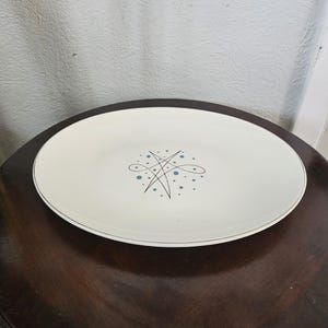 May include: An oval, cream-colored serving platter with a thin silver rim. The center features a stylized design of interconnected lines and small blue dots. The platter is set on a dark brown wooden table.