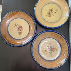 May include: Three round ceramic plates with a speckled tan center and a dark blue rim. Each plate features a unique floral or botanical illustration in the middle, including a red rose, a cluster of berries, and a bouquet of small flowers.