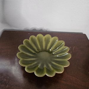 May include: A green, flower-shaped ceramic dish with a scalloped edge. The dish has a glossy finish and a central depression. The color is a muted olive green. The dish is likely for serving small items or as a decorative piece.