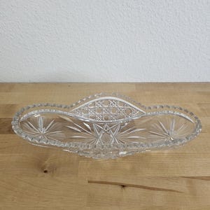 May include: An elongated clear glass dish featuring intricate cut glass patterns, including starburst motifs on the ends and a central lattice design. The rim of the dish has a scalloped edge, and the overall construction is transparent glass.