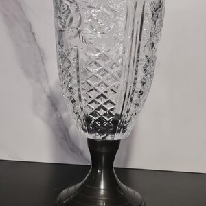 May include: A clear glass vase with a diamond pattern and floral relief, mounted on a dark metal base. The vase has a wide, flared top and a detailed, textured surface. The base is a dark, metallic color.