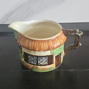May include: A ceramic creamer shaped like a cottage. The creamer features a thatched roof, windows, and a door. The handle is designed to look like a tree branch. The creamer is painted with brown, green, and beige colors.
