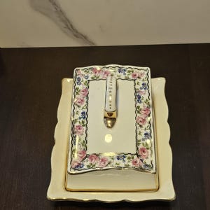 May include: A rectangular ceramic butter dish with a lid featuring a floral pattern of pink roses and blue flowers, accented by a black and white checkered border. The lid has a central handle with gold trim, and the base has scalloped edges.