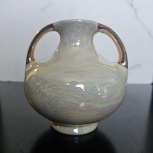 May include: A small, round ceramic vase with two arched handles on the sides. The surface features a pearlescent, iridescent finish with swirling patterns of light gray, cream, and subtle rainbow hues, complemented by metallic gold-toned handles.