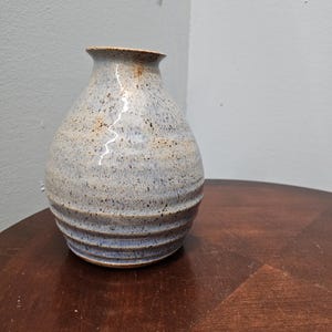 May include: A handmade ceramic vase with a speckled blue and tan glaze. The vase has a rounded body with horizontal ridges and a narrow neck. The vase is displayed on a dark brown wooden surface.