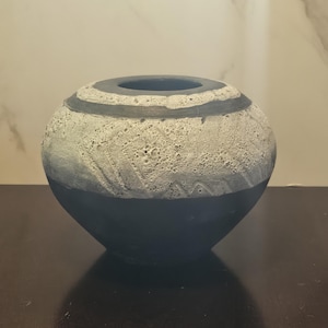 May include: A round, decorative vase with a textured, light gray upper half and a dark gray lower half. A dark gray band encircles the opening. The vase sits on a dark brown surface.