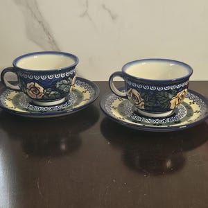 May include: Two matching ceramic teacups and saucers. The cups and saucers are navy blue with a floral design featuring pink and yellow roses. The rims of the cups are light blue. The saucers have a yellow and green border.