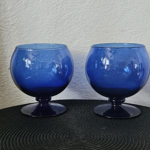 May include: Two cobalt blue glass goblets with a rounded bowl and a short stem. The glasses are displayed on a black textured surface. The goblets have a vintage aesthetic.