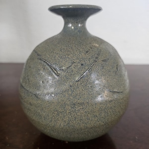 May include: A small, round, speckled ceramic vase in shades of gray and green. The vase has a narrow neck and a flared rim. Decorative lines are etched into the surface, adding texture. The vase is suitable for displaying small flowers or as a decorative accent.
