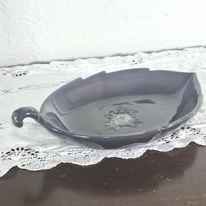 May include: A dark gray ceramic leaf-shaped serving dish with a handle. The dish has a raised floral design in the center and sits on a white lace tablecloth. The handle is curved and has a small decorative element.