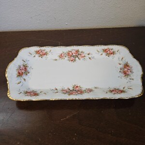 May include: A rectangular, white porcelain serving tray with a scalloped edge and gold trim. The tray is decorated with clusters of pink and peach roses and green leaves. A classic piece for serving appetizers or desserts.