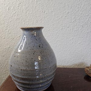May include: A handmade ceramic vase with a speckled blue-gray glaze. The vase has a rounded body with horizontal ridges near the base and a narrow neck. The rim is a contrasting brown color. This decorative vase is suitable for home decor.
