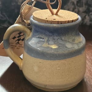 May include: A handmade ceramic jar with a cork lid and leather loop. The jar is beige with a blue band and handle. A tag is attached with a leather string. The jar is likely for kitchen or home decor.
