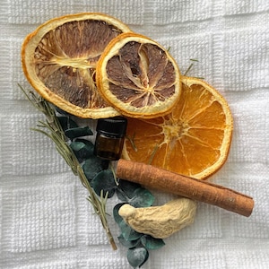 Puede incluir: Un arreglo de rodajas de naranja secas, una pequeña botella marrón de aceite esencial, una rama de canela, jengibre y ramitas de romero y eucalipto sobre una superficie blanca texturizada. Las rodajas de naranja son de color naranja vibrante.
