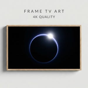 Può includere: Stampa incorniciata con un'immagine celeste di un'eclissi solare. L'opera mostra un sole bianco brillante circondato da un anello blu su uno sfondo nero. Il testo "FRAME TV ART" e "4K QUALITY" è visualizzato in alto.