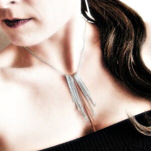 May include: A silver necklace with multiple vertical bar pendants and matching earrings. The jewelry is worn with a black strapless top. The woman has dark hair and is looking down.