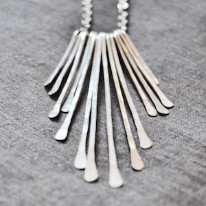 May include: A silver necklace with a fan-shaped pendant made of multiple thin, hammered metal strips.