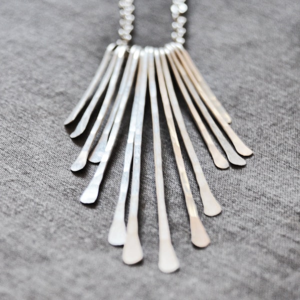 May include: A silver necklace with a fan-shaped pendant made of multiple thin, hammered metal strips.