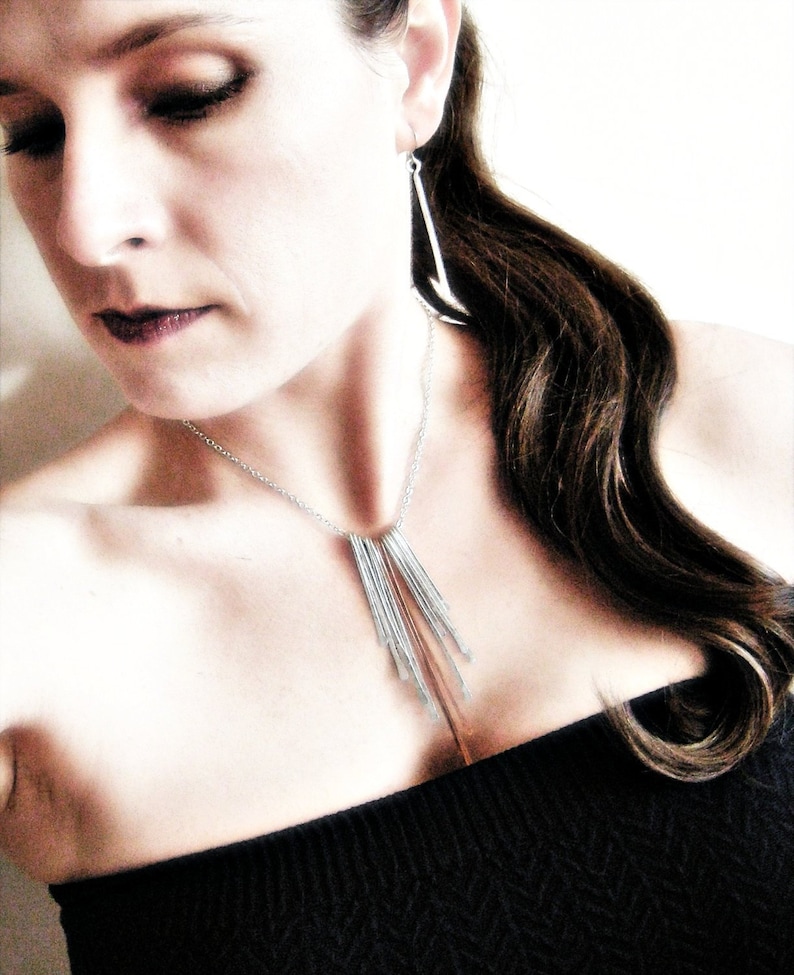 May include: A silver necklace with multiple vertical bar pendants and matching earrings. The jewelry is worn with a black strapless top. The woman has dark hair and is looking down.