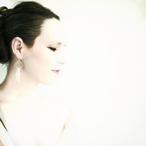 May include: A woman with dark brown hair styled in a bun, wearing silver dangle earrings with a circle design. The background is a soft white.