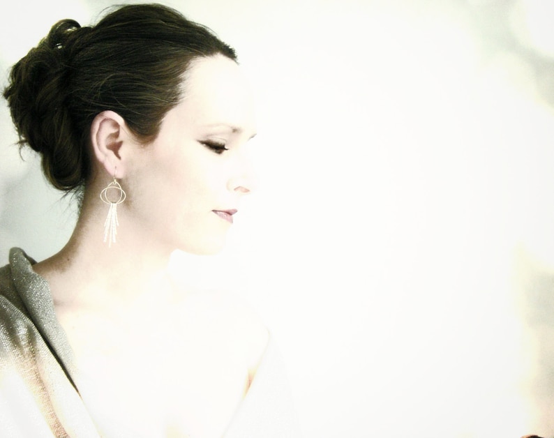 May include: A woman with dark brown hair styled in a bun, wearing silver dangle earrings with a circle design. The background is a soft white.